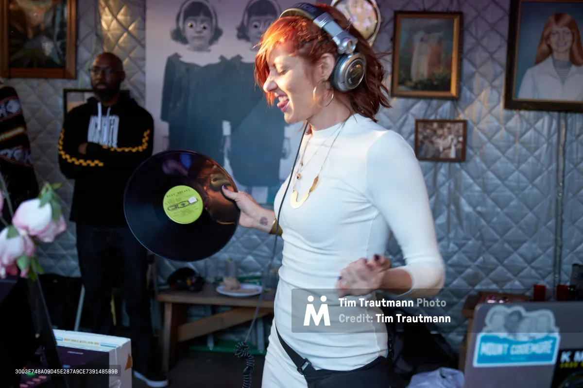DJ Doctor Dakar commands the booth at White Owl Social Club, her flame-red hair catching golden light as she grips a vintage vinyl record with infectious enthusiasm. The intimate Portland venue pulses with afternoon energy, its walls adorned with eclectic artwork creating a bohemian backdrop. Her pure joy radiates through professional headphones and crisp white attire, embodying the authentic spirit of vinyl culture in the Pacific Northwest music scene.
