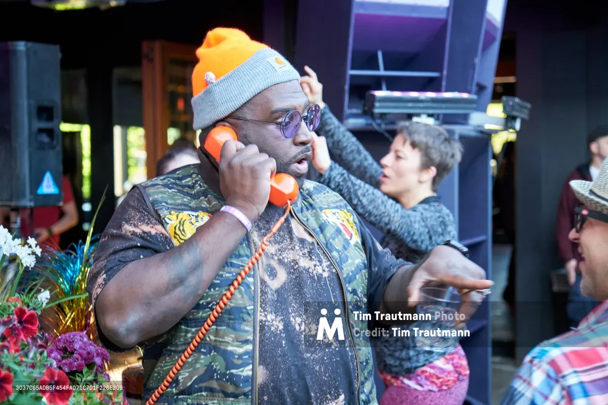 A man in military-style clothing and purple sunglasses holds a vintage orange rotary phone to his ear while speaking animatedly at Your Sunday Best's season opener. Behind him, partygoers mingle in the warm, ambient lighting of White Owl Social Club, with vibrant flowers adding splashes of color to the foreground. The scene captures the playful, nostalgic atmosphere of Portland's beloved summer day party series.
