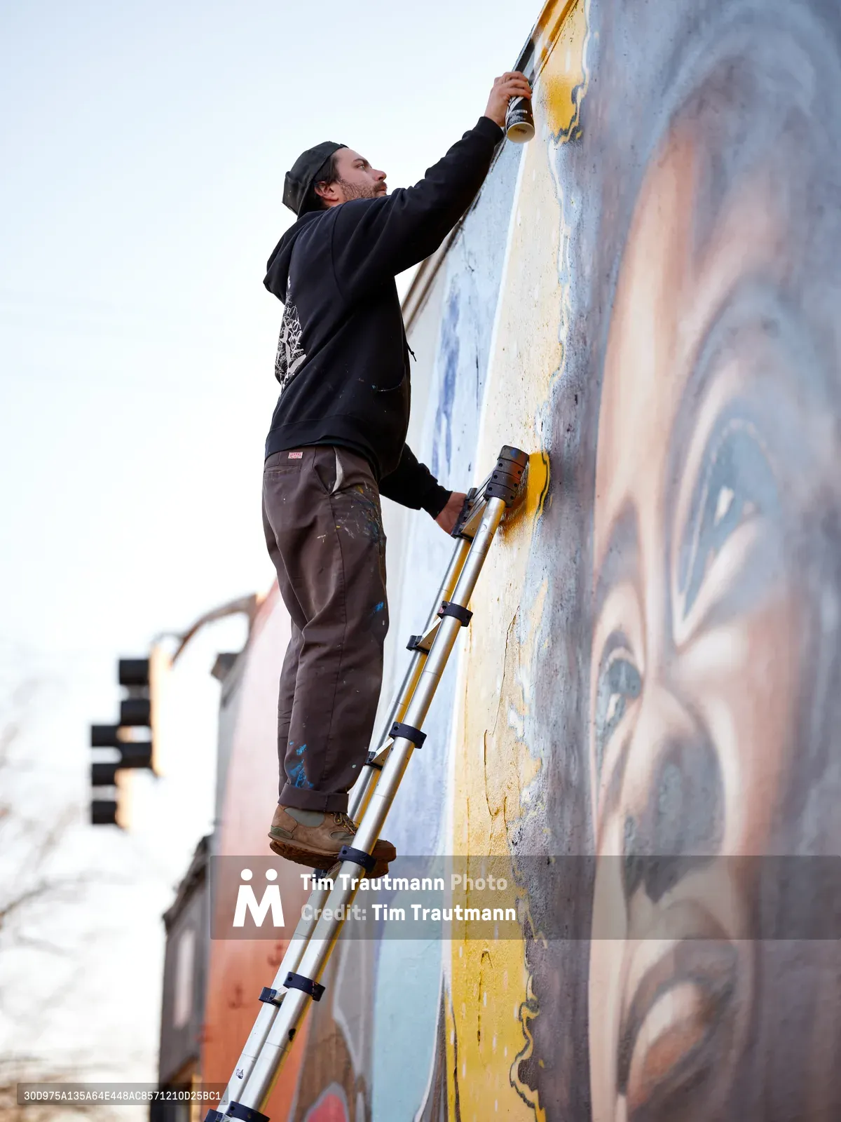 Travis Fields, known as campographic, stretches upward on an aluminum ladder to apply paint to a vibrant mural depicting poet Amanda Gorman. The street artist works against weathered concrete in Portland's Alberta Arts District, his dark hoodie and paint-stained work pants capturing the dedication of public art creation. Soft afternoon light illuminates the emerging portrait, revealing layers of blue, yellow, and flesh tones that bring the celebrated poet's likeness to life on the urban canvas.