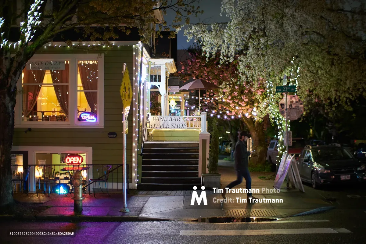 A solitary figure moves through the twilight streets of Portland's Nob Hill district, passing illuminated storefronts including a wine bar and bottle shop. Warm golden light spills from shop windows while delicate string lights trace the building's architectural lines, creating an intimate urban atmosphere. Spring blossoms on nearby trees add a seasonal softness to the hardscape, while purple neon signs cast colorful reflections on the wet pavement.