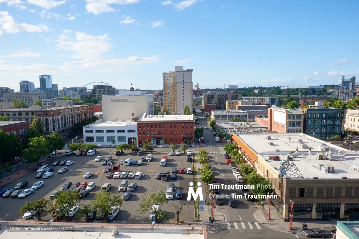 An elevated view captures the bustling intersection of Portland's historic Chinatown district, where a sun-drenched parking lot anchors the urban composition. Mid-rise buildings in varied architectural styles create a textured skyline against wispy white clouds, while the Kalberer building stands prominently among mixed-use structures. The afternoon light bathes the scene in warm tones, revealing the neighborhood's blend of residential towers, commercial buildings, and tree-lined streets that define this evolving urban landscape.