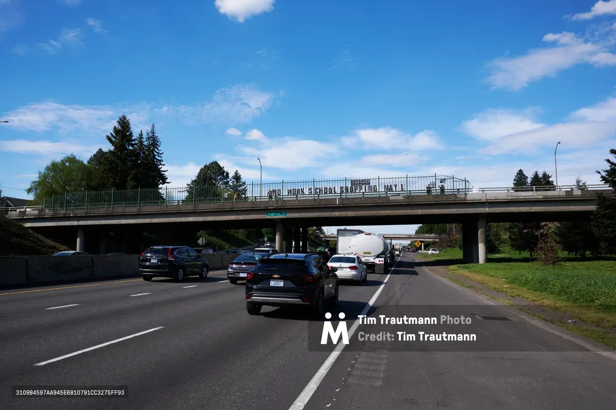 Activists display a protest banner reading 'No Work School Shopping May 1st' from a pedestrian overpass spanning Interstate 5 in North Portland's Humboldt neighborhood. The concrete bridge frames the banner against a brilliant blue spring sky dotted with white cumulus clouds, while morning traffic flows steadily beneath. Towering evergreens and mixed deciduous trees create a verdant backdrop, their fresh green foliage suggesting the renewal of spring organizing season.