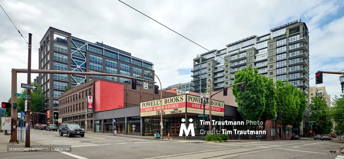 The iconic Powell's City of Books stands eerily quiet at the intersection of West Burnside Street in Portland's Pearl District during the COVID-19 pandemic lockdown of May 2020. The legendary bookstore's familiar corner location, with its distinctive red and cream facade, sits empty beneath overcast skies while modern residential towers loom behind. Traffic lights cast their colored glow over nearly deserted streets, creating a haunting portrait of urban isolation during one of the city's most challenging periods.
