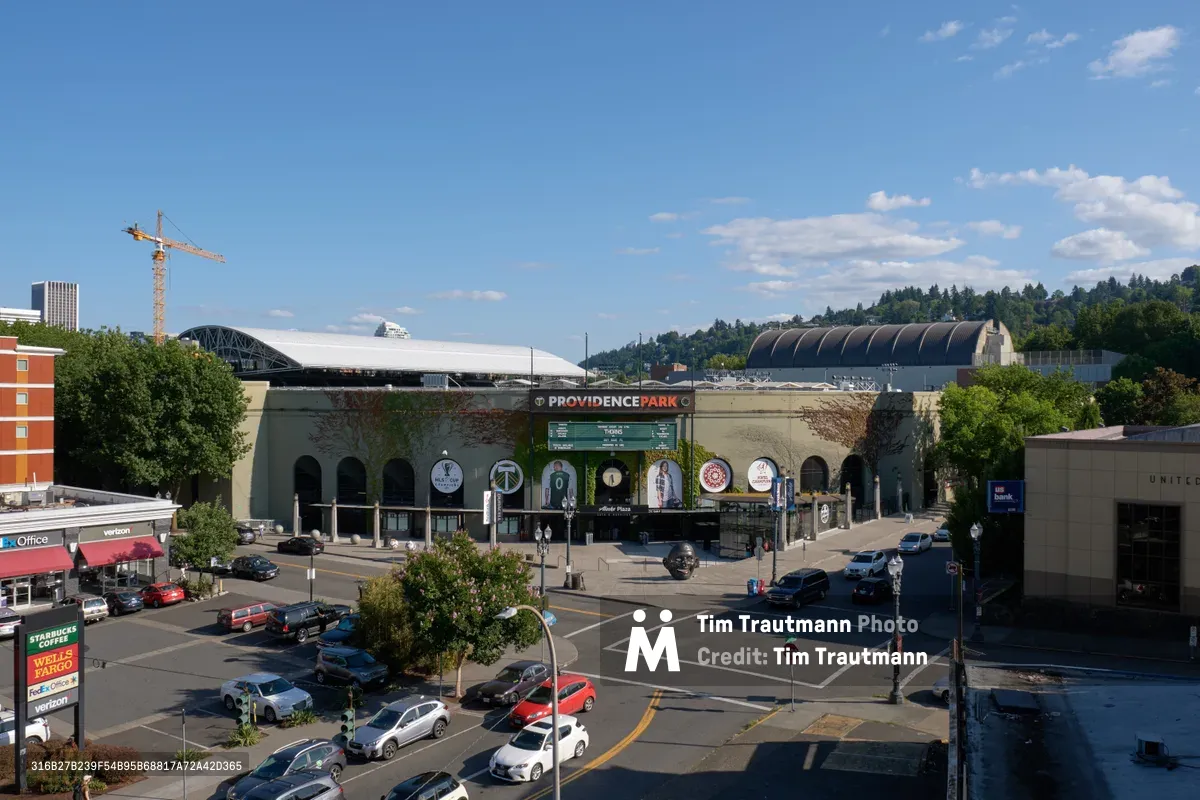 An elevated view captures Providence Park's distinctive ivy-covered facade beneath a brilliant azure sky dotted with cotton-white clouds. The historic stadium anchors a bustling Goose Hollow intersection where urban traffic flows around the soccer venue's weathered concrete exterior. Construction cranes pierce the horizon beyond the arena, while forested hills create a verdant backdrop to Portland's evolving cityscape.