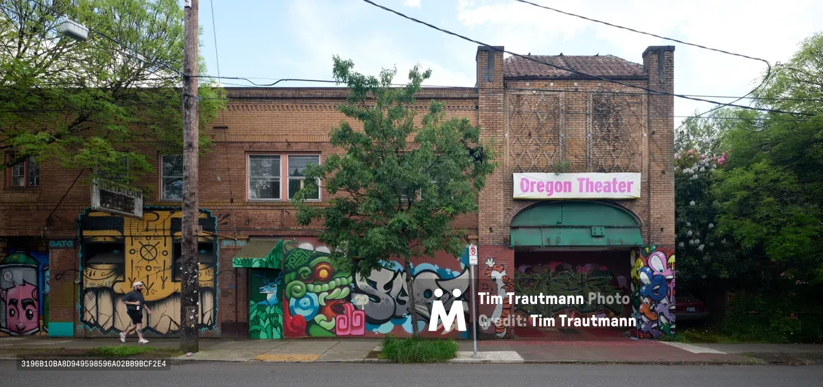 Vibrant graffiti and street art murals transform the weathered brick facade of the former Oregon Theater on Southeast Division Street in Portland's Richmond neighborhood. The eclectic artwork spans the building's ground level, featuring bold characters, geometric patterns, and colorful designs that breathe new life into this historic entertainment venue. Lush spring foliage frames the scene while power lines stretch overhead, capturing the organic intersection of urban decay and artistic renewal that defines Portland's evolving streetscape.
