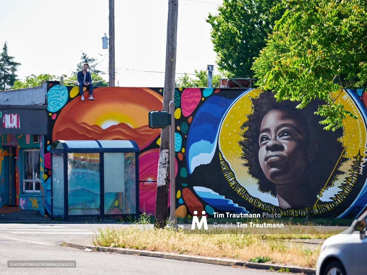 Travis Fields sits triumphantly atop a weathered rooftop, having just completed his vibrant mural honoring poet Amanda Gorman in Portland's Alberta district. The sprawling street art below bursts with warm sunset gradients, swirling organic shapes, and Gorman's portrait rendered with a golden halo against celestial blues. Dappled afternoon light filters through verdant tree canopy, casting the scene in the golden hour glow of creative accomplishment.