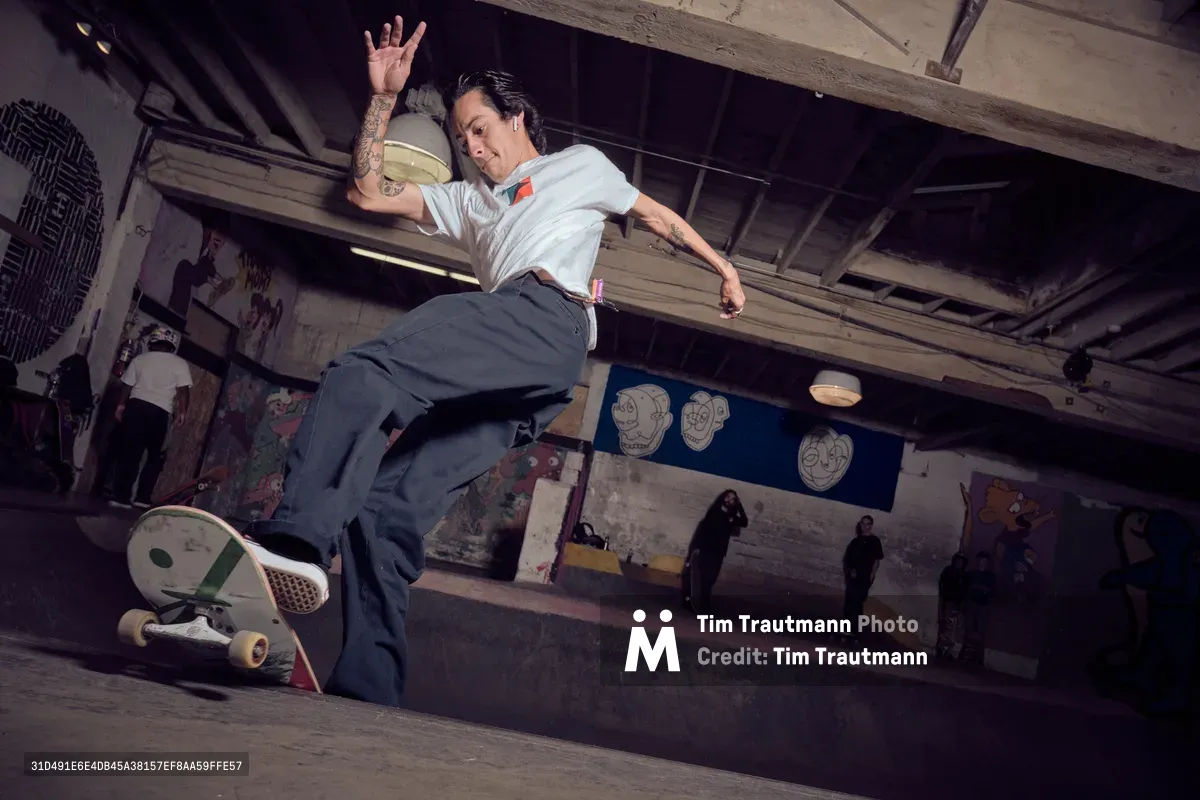 A skateboarder launches into weightless suspension above the concrete bowl at Commonwealth Skateboarding in Portland's Buckman neighborhood, arms spread wide against the industrial ceiling's exposed beams. The underground skate park's graffitied walls and scattered onlookers create a gritty urban cathedral where street culture thrives beneath fluorescent light. The moment captures pure athletic poetry—body extended in flight, board trailing below, while the venue's raw concrete and tagged surfaces frame this expression of underground artistry.