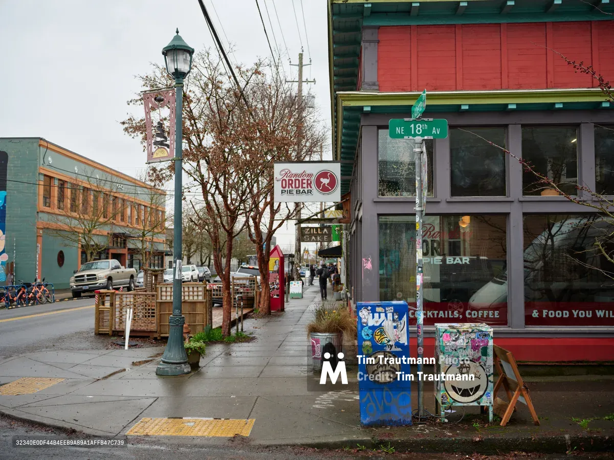 A wet sidewalk scene captures the vibrant character of Portland's Alberta Street, where the Random Order Pie Bar anchors a block of colorful storefronts and street art. Overcast skies cast even light across the rain-dampened pavement, while vintage lampposts and power lines create urban rhythms above. Sticker-covered utility boxes and weathered signage speak to the neighborhood's artistic spirit and community energy.