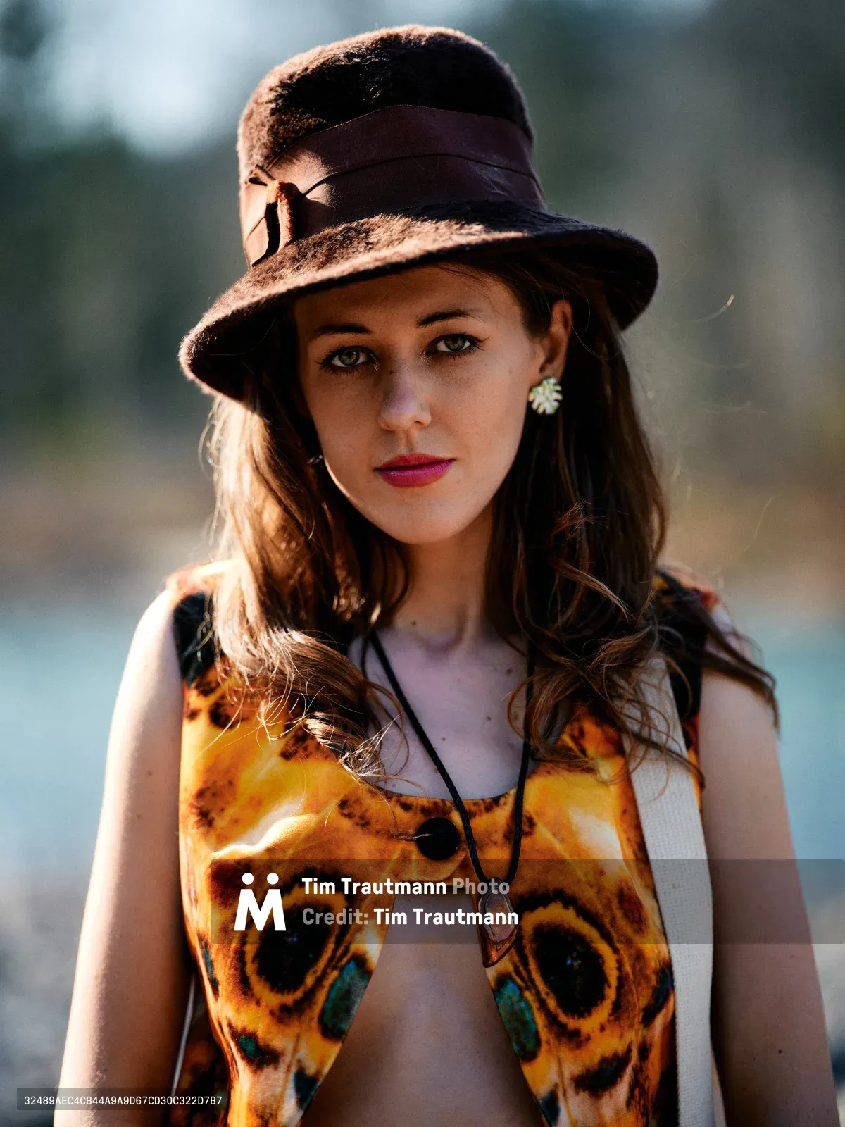 A young woman with flowing chestnut hair poses confidently along Oregon's Floodplain Trail, wearing a striking butterfly-patterned top and vintage brown fedora. Golden hour light creates dramatic rim lighting on her hair and hat while casting warm shadows across her face. Her bohemian styling includes flower earrings and layered necklaces, embodying free-spirited outdoor fashion against the softly blurred riverbed backdrop of Multnomah County's scenic landscape.