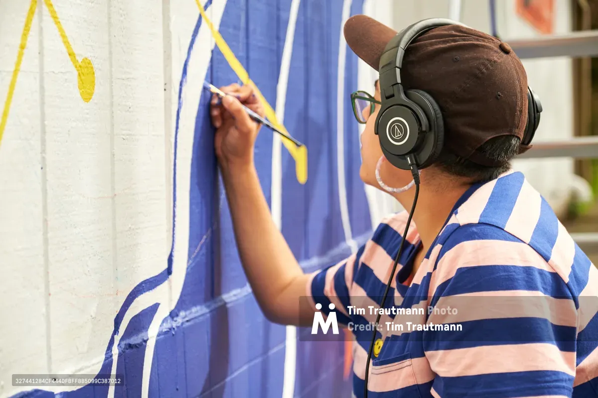 Maria Rodriguez, known as Sparkykneecap, works on her commissioned mural "Let's Talk" at Open Signal in Portland's Eliot neighborhood. The Mexican-American artist wears headphones and a blue striped shirt while painting vibrant geometric shapes in yellow and blue on a white wall. Her careful brushwork brings themes of identity and cultural connection to life through bold, playful forms that invite community dialogue.