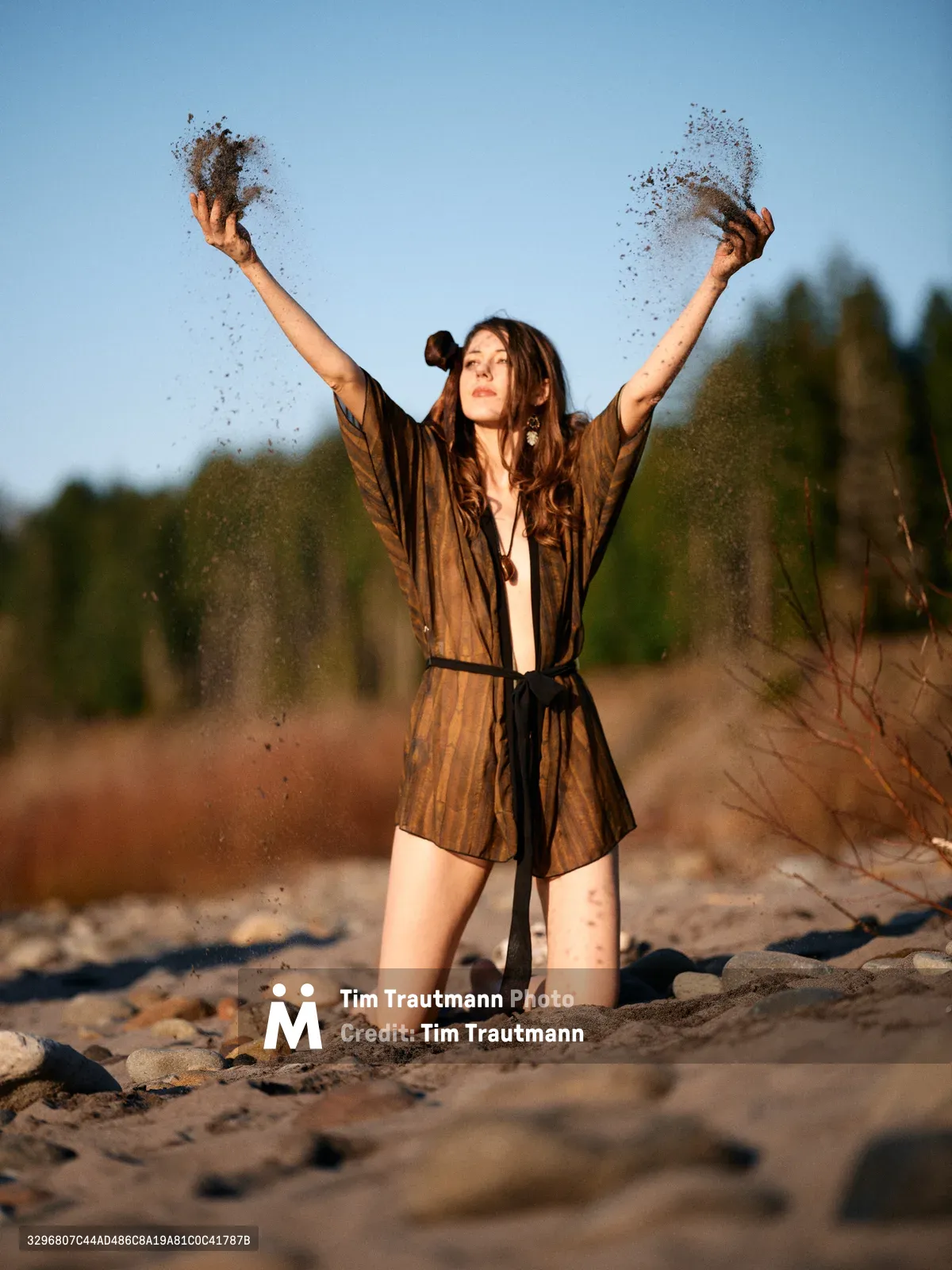 A young woman in an earth-toned robe kneels on the sandy riverbed along Oregon's Floodplain Trail, arms raised triumphantly as fine sand cascades from her open palms. The golden hour light bathes the scene in warm amber tones, while the blurred forest backdrop creates intimate depth. Her expression radiates pure joy and liberation, capturing a moment of spontaneous celebration amidst the natural riverside landscape of Multnomah County.