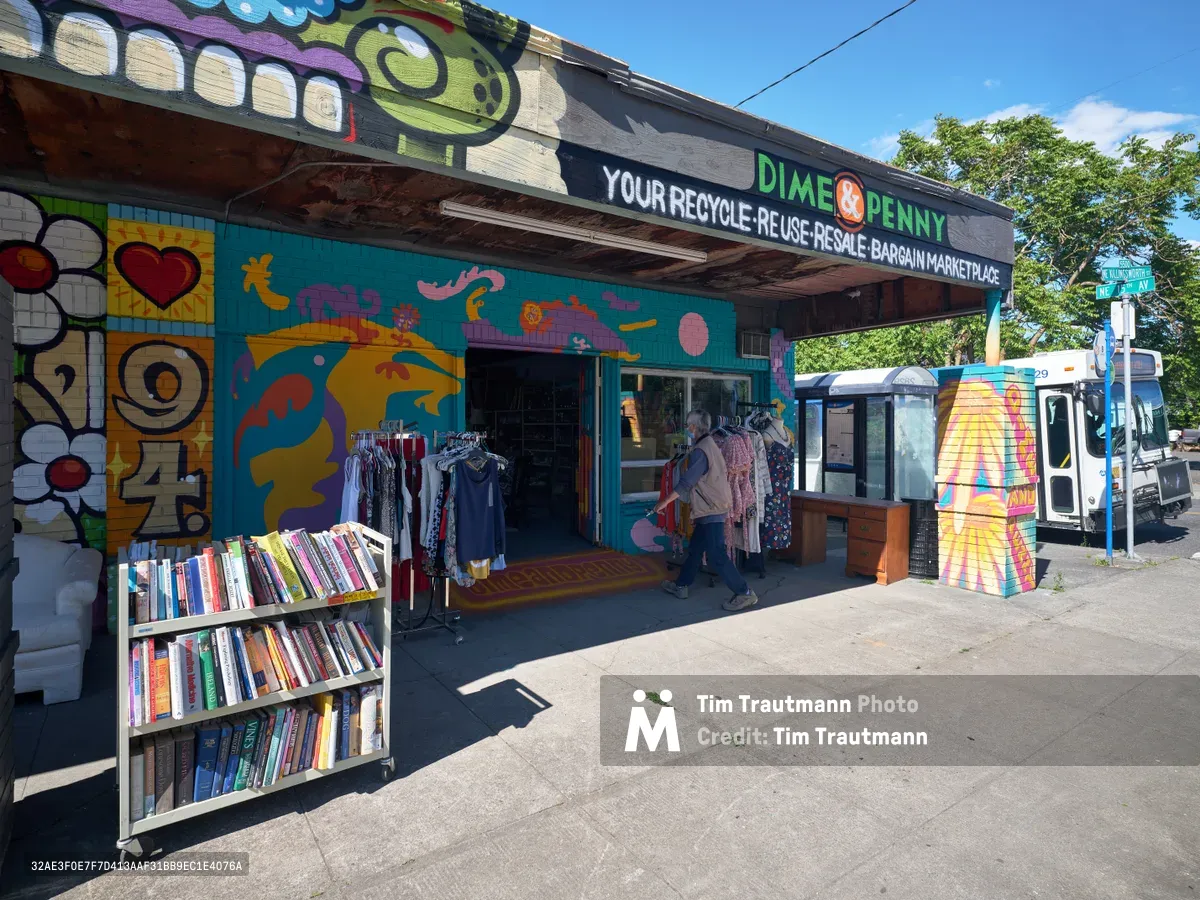 The vibrant Dime & Penny thrift store on Northeast 15th and Killingsworth displays its sustainable mission through bold murals and outdoor book displays. Afternoon sunlight illuminates the turquoise storefront adorned with colorful graffiti-style artwork featuring floating whales and abstract forms. A customer browses clothing racks beneath the covered entrance while rainbow-spined books create an inviting streetside library, embodying Portland's commitment to reuse and community exchange.