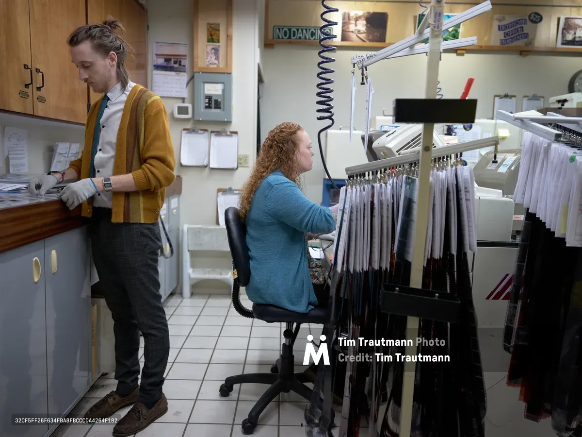Two film lab technicians work in concentrated silence within the intimate confines of Blue Moon Camera & Machine in Portland's Saint Johns neighborhood. A woman with curly hair in a teal sweater sits at a processing station surrounded by hanging film strips, while her colleague in a mustard cardigan examines prints at the counter. The cramped workspace pulses with the quiet intensity of analog craftsmanship, fluorescent light casting clinical precision over rows of negatives and the accumulated ephemera of photographic tradition.