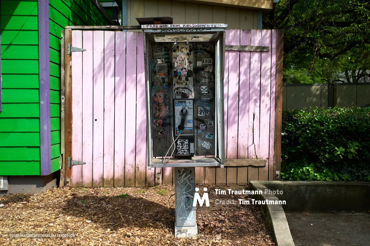A weathered payphone stands as a working relic of communication past, its pink wooden shutters opened to reveal a graffiti-covered interior densely packed with stickers and street art. Surrounded by urban debris with wood chips scattered at its base, the functional phone creates a stark contrast against the vibrant green siding of a nearby building. Dappled sunlight filters through overhead foliage, casting gentle shadows across this active piece of telecommunications infrastructure in Portland's Eliot neighborhood.