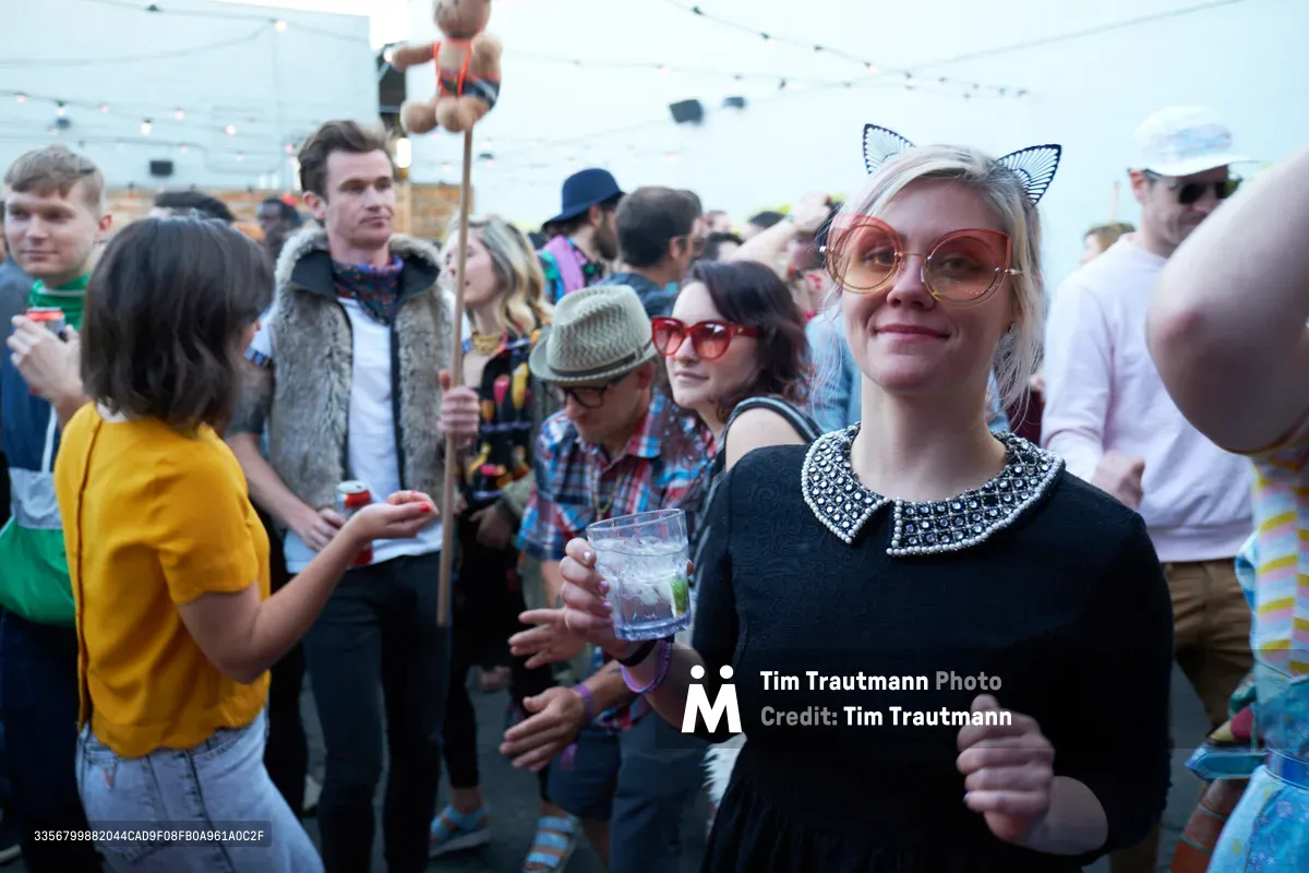 A blonde woman in a black dress with pearl collar and whimsical cat ear headband radiates joy while holding a cocktail at Your Sunday Best's season opener. The crowded rooftop gathering at White Owl Social Club buzzes with eclectic festival fashion, from vintage fur vests to yellow retro tops. Warm afternoon light bathes the diverse crowd of young adults mingling with drinks against the backdrop of Portland's urban skyline.