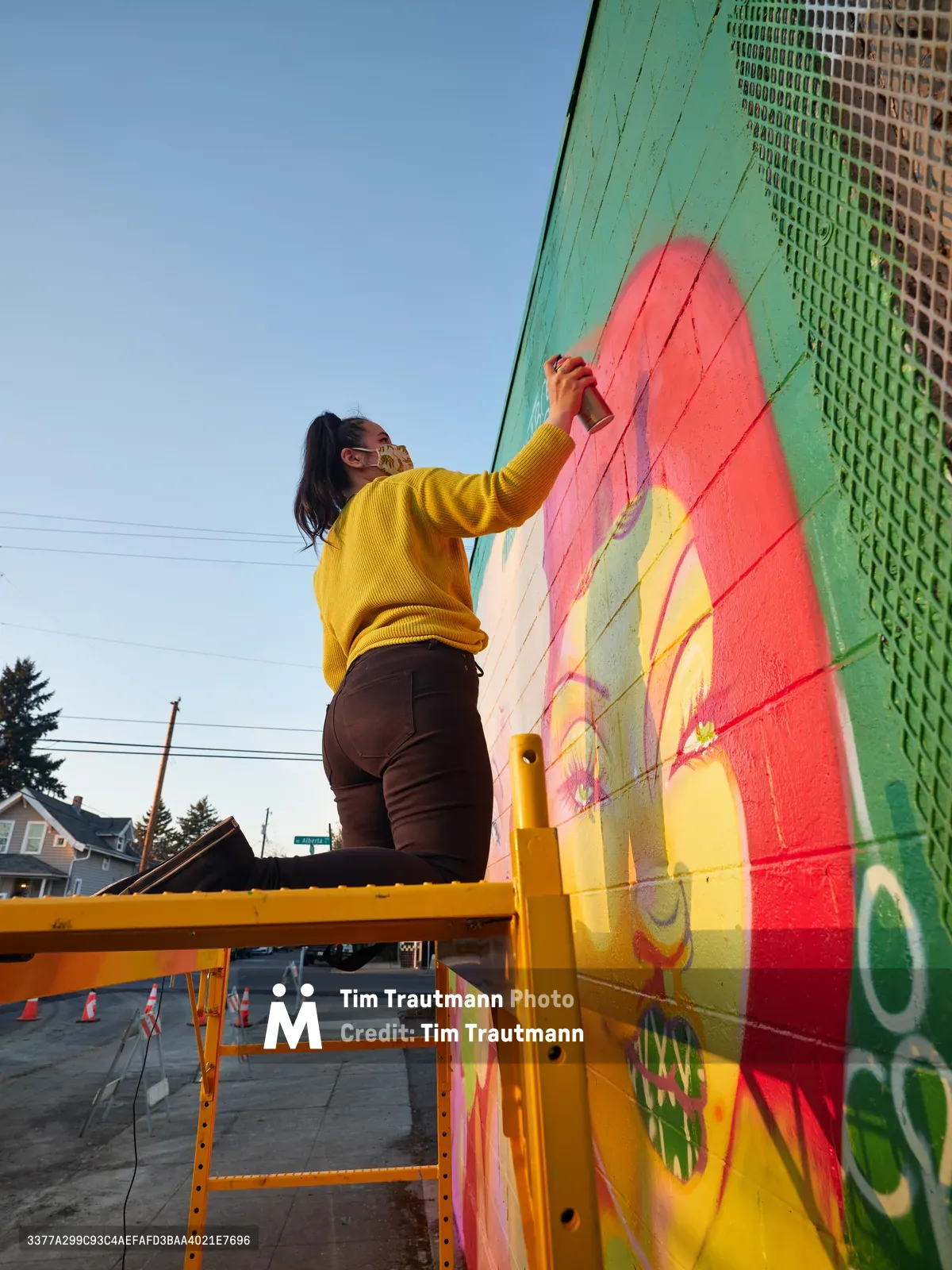 Multidisciplinary artist Villatose works from a yellow scissor lift, adding bold coral and yellow brushstrokes to her mural 'Jardín Cubana' featuring three female figures on Northeast Alberta Street in Portland's arts district. The late afternoon light bathes the scene as she meticulously applies paint to the mint green wall, her mustard sweater and dark pants creating a striking silhouette against the clear Oregon sky. Power lines and residential rooftops frame the background of this quintessential Portland neighborhood street art scene.