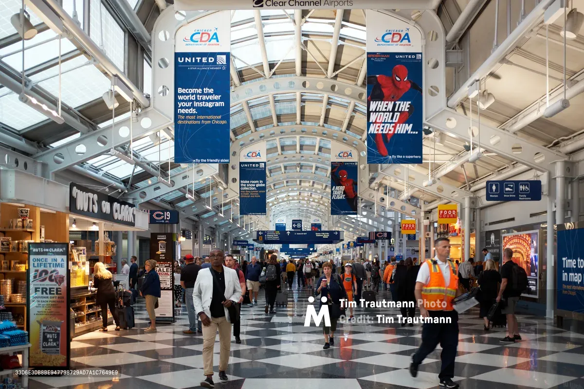 The soaring glass-and-steel concourse of Terminal C at Chicago O'Hare International Airport pulses with constant motion as travelers navigate beneath a dramatic barrel-vaulted ceiling. Natural light streams through skylights, illuminating the polished checkerboard floor where passengers in business attire mingle with airport workers in bright orange safety vests. Overhead, suspended advertising banners for airlines and travel destinations create a commercial canopy above the steady flow of departing and arriving travelers.