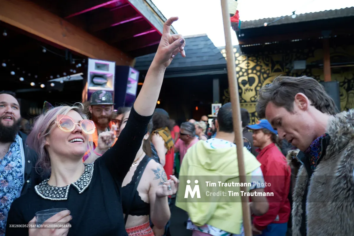 A jubilant woman with platinum blonde hair and round glasses throws her arm skyward in pure celebration, her face radiating joy as she dances among the crowd at White Owl Social Club's outdoor summer party. The warm afternoon light filters through the venue's industrial architecture, illuminating her black vintage-style dress and the diverse gathering of revelers around her. String lights and exposed beams frame the scene, while fellow dancers in colorful attire create a vibrant backdrop of community and connection.
