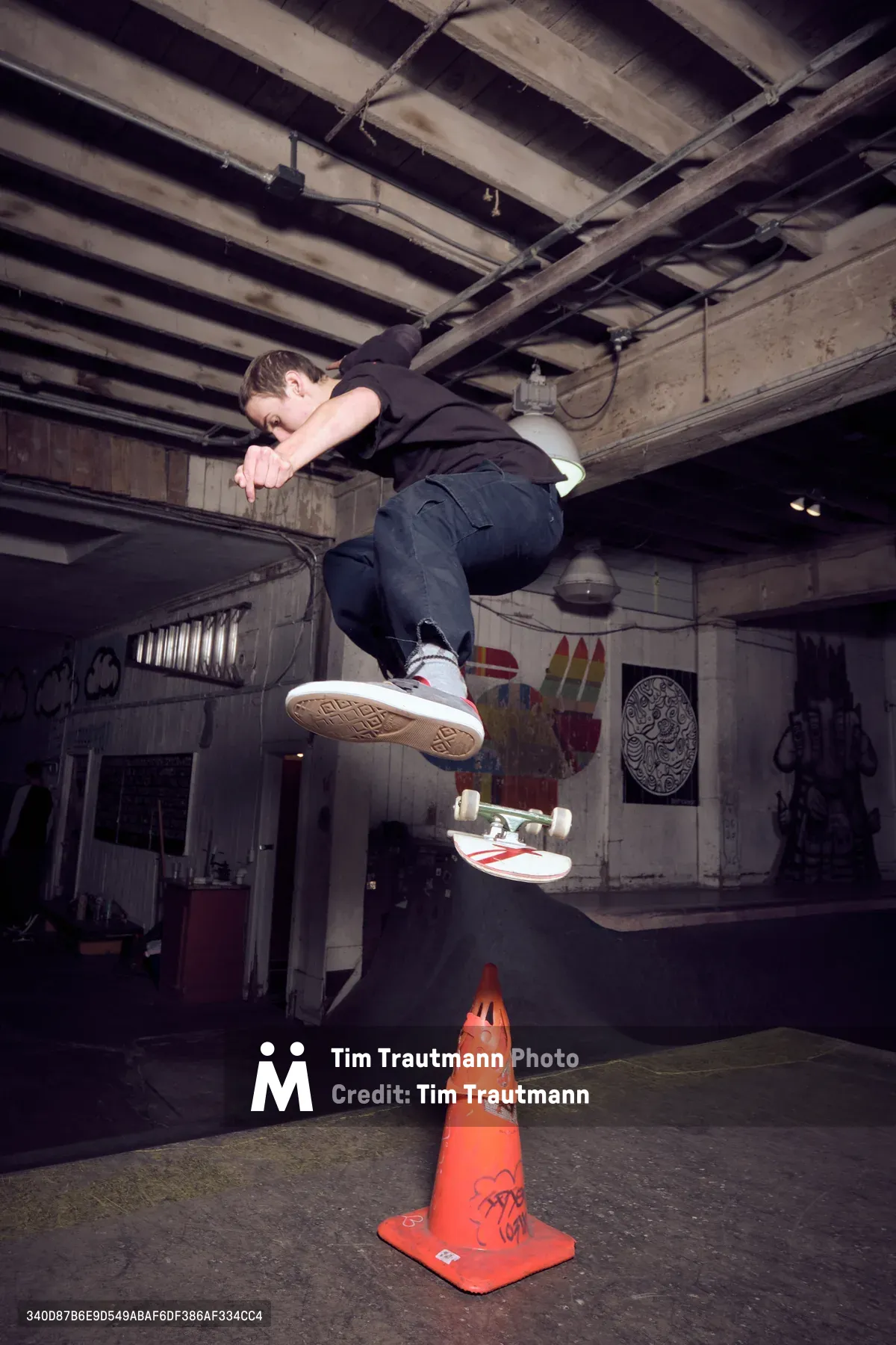 A young skateboarder launches into an aerial maneuver above an orange traffic cone at the dimly lit Commonwealth Skateboarding in Portland's Buckman neighborhood. The exposed wooden ceiling beams and graffitied concrete walls create an authentic underground atmosphere, while warm ambient lighting illuminates the skater mid-flight. Street art and skating culture merge in this intimate editorial moment captured in one of Portland's iconic skate shops.