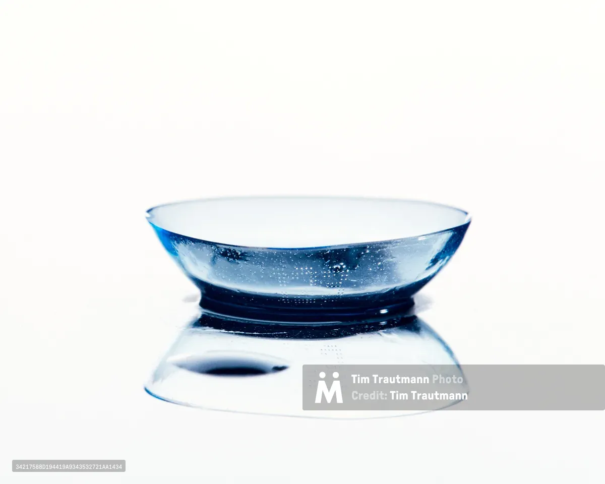 A single contact lens rests delicately on a pristine reflective surface, its transparent form capturing light and creating a mesmerizing display of refraction. Tiny bubbles suspended within the hydrogel material sparkle like captured starlight, while the lens's gentle curve creates a perfect mirror reflection beneath. The minimalist composition against a pure white background transforms this everyday object into an elegant study of transparency and optical precision.