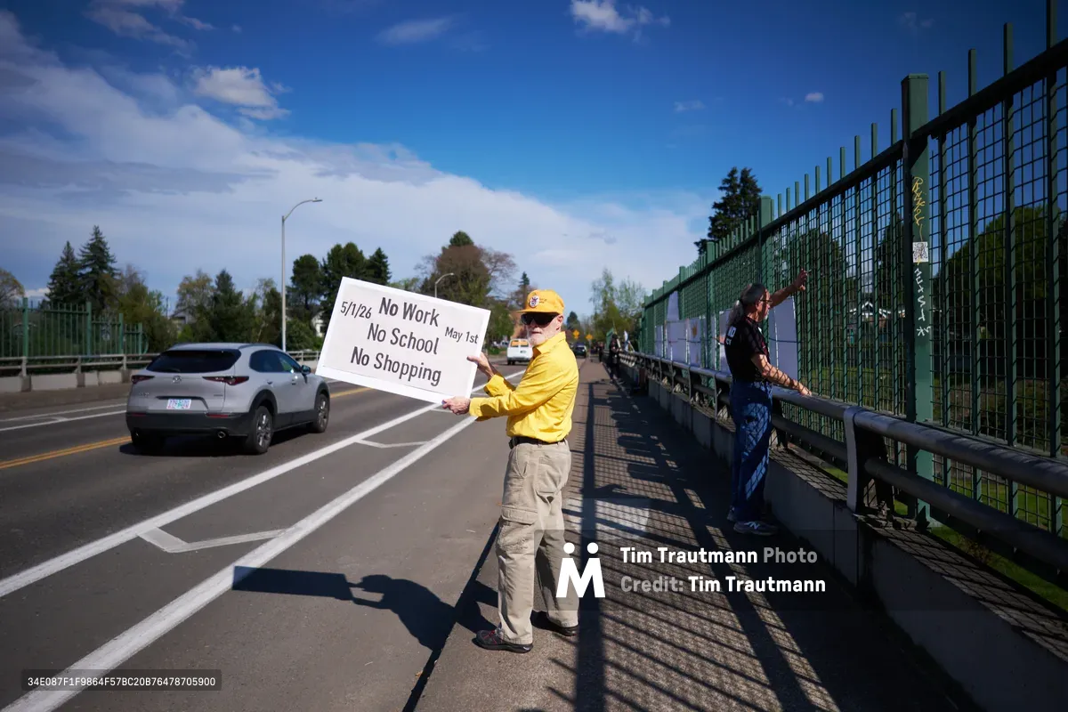 A lone activist in bright yellow jacket and cap stands on the North Skidmore Street overpass above Interstate 5 in North Portland, holding a handwritten protest sign calling for a May 1st General Strike. The demonstrator displays a banner reading "5/1/20 No Work No School No Shopping May 1st" while another person in dark clothing moves along the green metal fence. Afternoon sunlight casts sharp shadows across the concrete walkway as traffic flows beneath on the busy freeway, creating a powerful juxtaposition between grassroots activism and urban infrastructure.