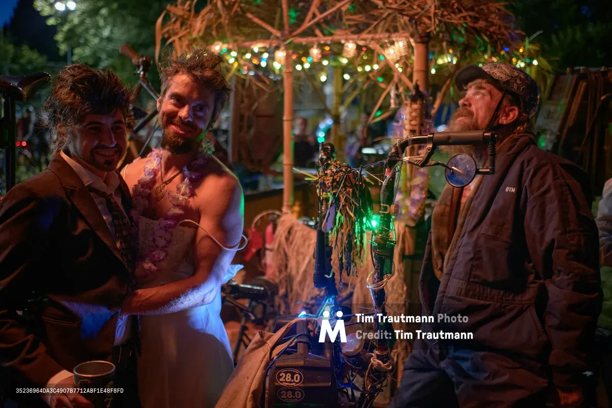 A joyous couple embraces amid the whimsical chaos of Portland's Drop Out Prom Ride, part of the city's annual Pedalpalooza cycling celebration. The bride, adorned in white with purple accents, beams alongside her groom while a goggle-wearing cyclist operates an elaborate steampunk contraption bathed in electric blue light. String lights twinkle overhead in Colonel Summers Community Garden, creating an enchanted atmosphere where alternative culture and community celebration converge.