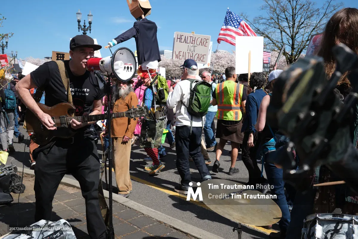 A guitarist in a black fedora and dark clothing performs passionately on Southwest Ankeny Street in Portland's historic Chinatown district, his instrument raised high as protesters march behind him carrying signs reading "HEALTH CARE NOT WEALTH CARE." The scene captures the intersection of music and activism under clear blue skies, with cherry blossoms framing the demonstration as drummers and marchers fill the street. American flags wave among the crowd while the musician's energetic performance adds a rebellious soundtrack to the political gathering.