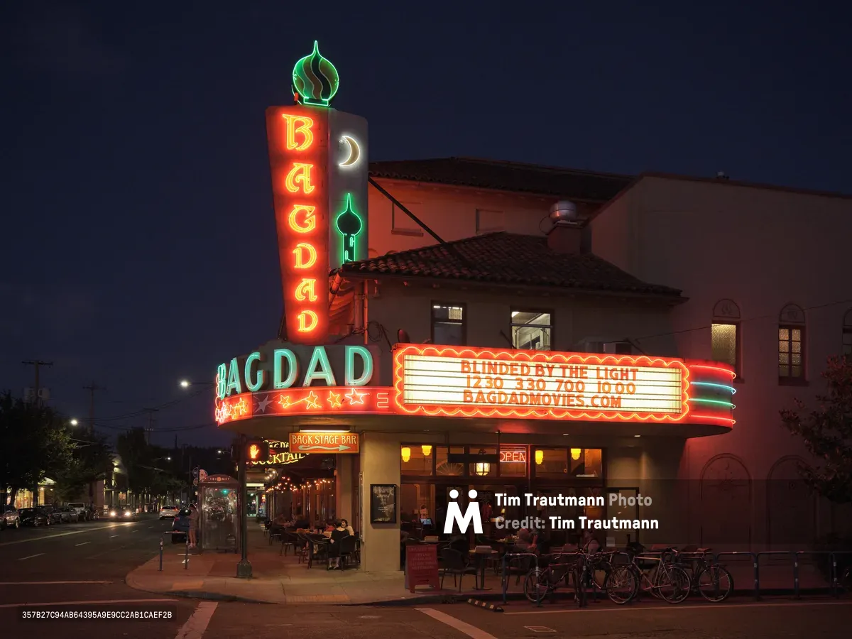 The historic Bagdad Theater & Pub radiates warmth against Portland's deepening twilight sky, its iconic vertical marquee blazing in red neon letters while the horizontal sign advertises showtimes. The Spanish Colonial Revival architecture frames the luminous entrance where patrons gather beneath the glowing canopy. Bicycles line the sidewalk and street lamps punctuate Southeast Hawthorne Boulevard, creating an intimate neighborhood cinema atmosphere in Richmond district.