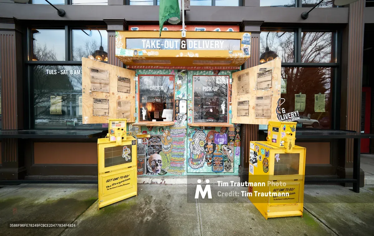 A vibrant street art-covered takeout window serves as the main entrance to Lottie and Zula's restaurant in Portland's Eliot neighborhood during the COVID-19 pandemic. Flanked by bright yellow Oregonian newspaper boxes, the colorful custom-built order window features psychedelic murals of skulls and cartoon characters, while handwritten menus on kraft paper frame the glass opening. The overcast afternoon light filters through bare winter branches, casting a subdued atmosphere over this adaptive pandemic streetscape.