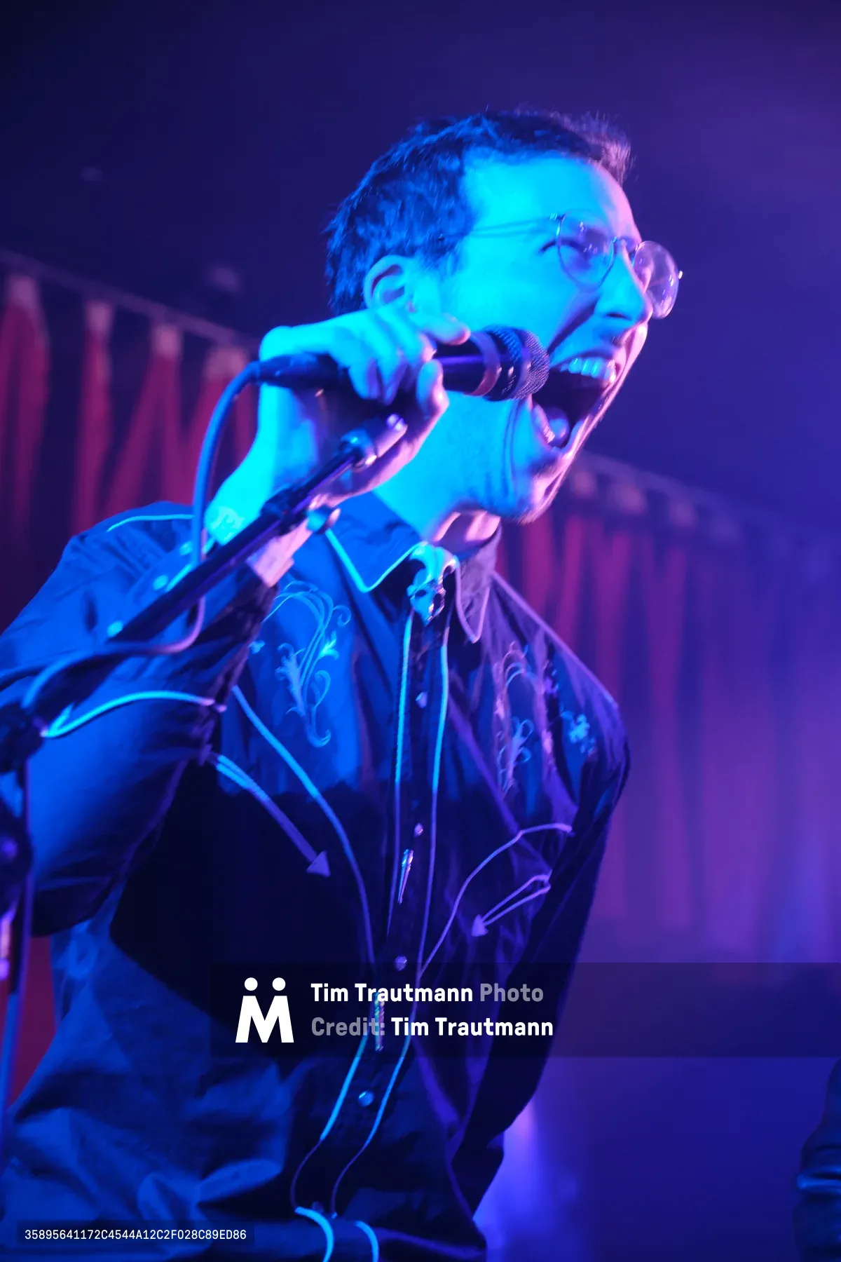 A vocalist from Roselit Bone commands the microphone under saturated blue and purple stage lighting at Dante's in Portland, Oregon. The performer's intense expression and wire-rimmed glasses catch the theatrical illumination as he leans into his delivery, creating a raw intimacy typical of the legendary venue's underground music scene. The moody lighting transforms the artist into a silhouette of electric energy against the club's shadowy interior. This captures the visceral connection between performer and audience that defines Portland's alternative music culture.