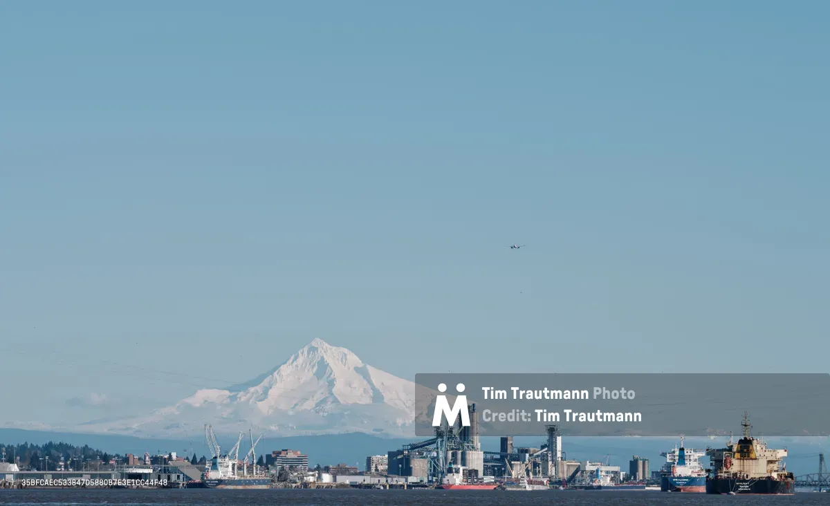 Mount Hood's snow-capped peak rises majestically beyond the industrial sprawl of the Port of Vancouver, Washington, as viewed from Kelley Point Park in Oregon. The crystalline morning light illuminates the mountain's glaciated slopes while cargo ships and port infrastructure create a striking juxtaposition of natural grandeur and maritime commerce. A small aircraft traces across the pale blue sky, adding scale to this panoramic vista where the Columbia River separates two states.