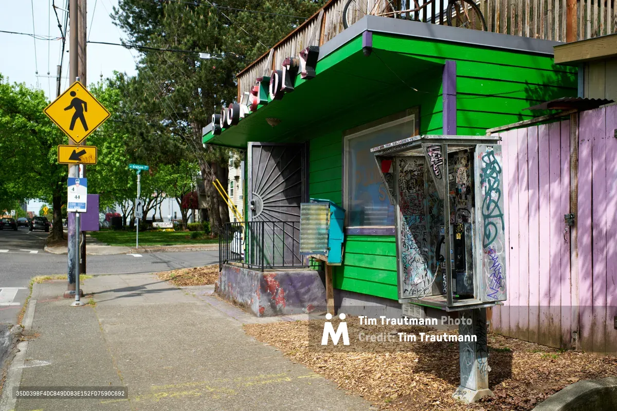 A weathered pay phone stands sentinel on a North Williams Avenue corner in Portland's Eliot neighborhood, its metal frame adorned with layers of colorful street art and stickers. The phone sits beside a vibrant green wooden building with purple trim, while pedestrian crossing signs and urban infrastructure frame the scene under diffused daylight. Wood chips scattered at the base and power lines overhead complete this quintessential portrait of Portland's eclectic streetscape.