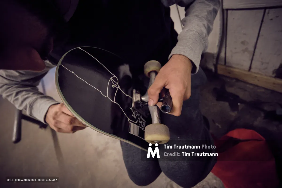 A skateboarder's weathered hands cradle a black skateboard deck adorned with intricate white line graphics, carefully adjusting the trucks and hardware in the warm, industrial atmosphere of Commonwealth Skateboarding in Portland's Buckman neighborhood. The intimate scene captures the ritualistic care between rider and board, with soft lighting illuminating the precise mechanical work. Red accents punctuate the shadowed workspace, while the concrete floor and utilitarian setting speak to the authentic culture of Southeast Portland's skateboarding community.