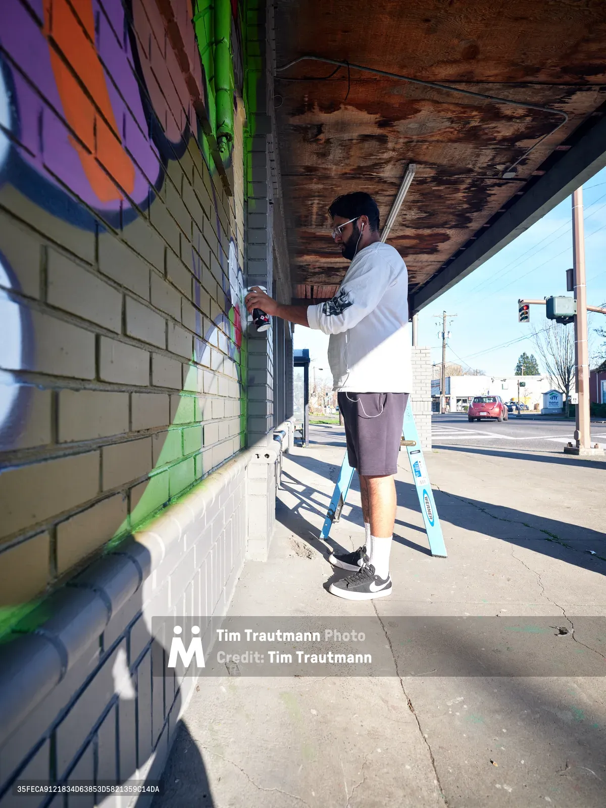 Muralist Heysus works methodically on a vibrant street art piece beneath the weathered overhang of a building at the intersection of Killingsworth and NE 15th Avenue in Portland's Alberta district. Afternoon sunlight streams across the concrete sidewalk, casting sharp shadows as the masked artist in white hoodie and shorts applies spray paint to transform the pale brick wall with bursts of purple, orange, and green. The urban canvas emerges at this bustling Northeast Portland corner, where residential streets meet artistic expression.