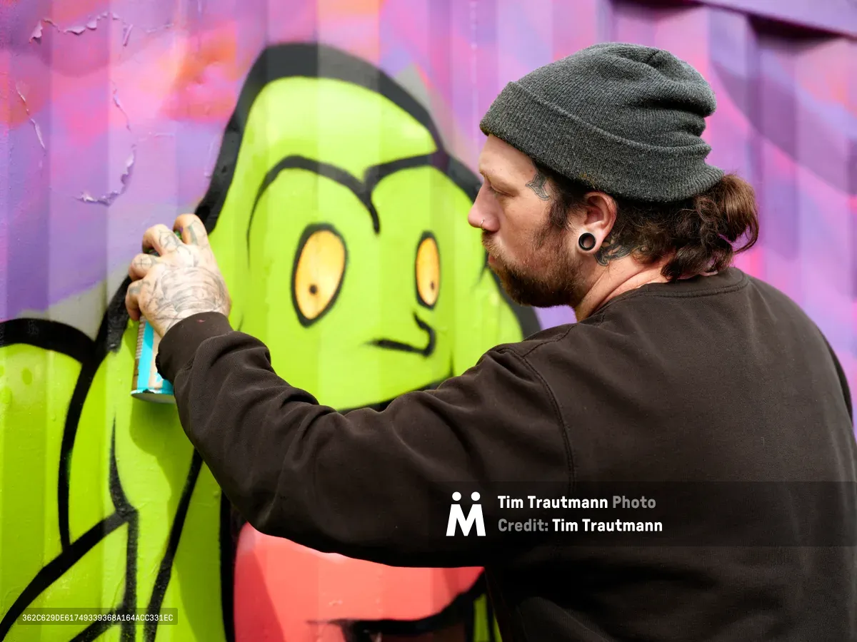 A bearded artist in a charcoal beanie and dark hoodie meticulously works on a bright lime green cartoon character against a vivid purple and pink backdrop. The intimate moment captures the focused concentration of street art creation, with the artist's tattooed hands delicately adding details to the whimsical figure's yellow eyes. The weathered urban wall provides texture and authenticity to this Portland neighborhood art scene.