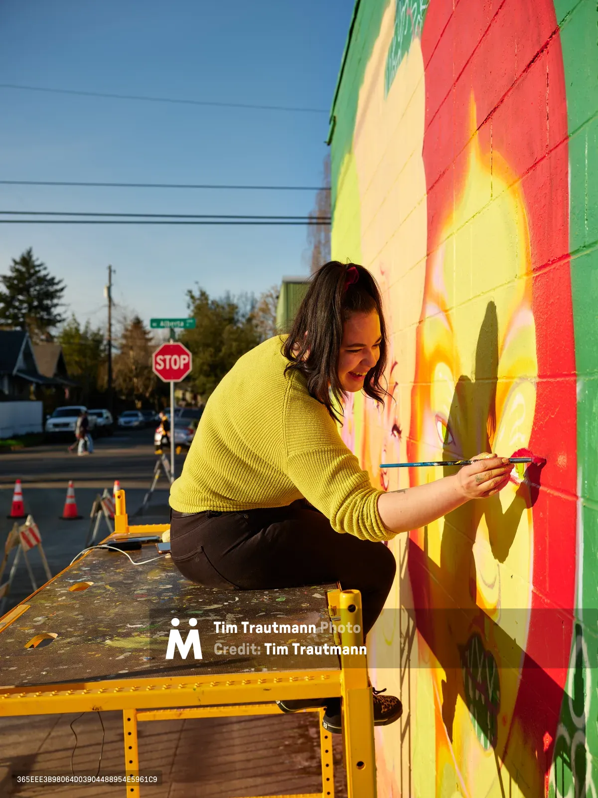 Multidisciplinary artist Vilatose works intently on her mural Jardín Cubana, depicting three female figures along Northeast Alberta Street in Portland's celebrated arts district. Golden hour light bathes the scene as she applies precise brushstrokes to the colorful wall, her yellow sweater echoing the warm tones of her artistic creation. The urban setting captures the creative energy of Portland's Alberta Arts District, where community murals transform neighborhood walls into canvases of expression.