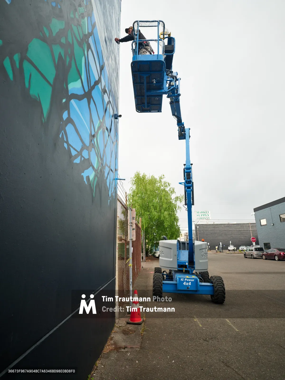 Artists Jeremy Nichols and Devin Finley work from the elevated platform of a blue cherry picker lift, applying vibrant green and blue paint to a large-scale mural on a concrete wall in Southeast Portland's Central Eastside district. The industrial setting features overcast skies and urban infrastructure, with the mechanical lift creating a dramatic vertical line against the sprawling street art composition. Safety cones mark the work zone while the artists concentrate on their collaborative public art project for the Portland Street Art Alliance.