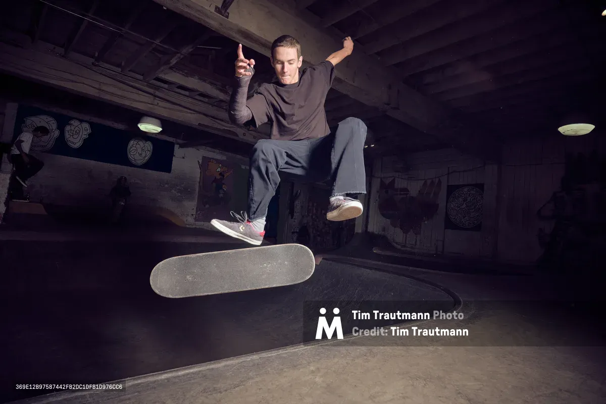 A skateboarder executes a dynamic flip trick above a concrete bowl at Commonwealth Skateboarding in Portland's Buckman neighborhood. The dramatic low-angle perspective captures the athlete suspended in mid-air, his board spinning beneath him as he extends both arms for balance. Moody artificial lighting illuminates the underground skate park's weathered concrete surfaces and graffitied walls, creating stark shadows that emphasize the intensity of the moment.