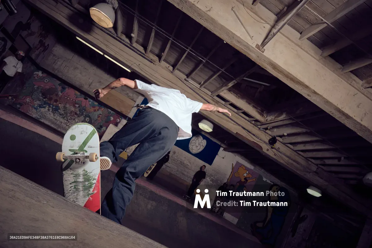 A skateboarder executes a dynamic aerial maneuver in the raw concrete basement of Commonwealth Skateboarding in Portland's Buckman neighborhood. The dramatic low-angle perspective captures the athlete mid-flight against exposed ceiling beams and weathered concrete pillars, while graffiti and street art provide colorful contrast to the industrial architecture. Moody lighting filters through the underground space, creating deep shadows that emphasize the gritty urban aesthetic of this Southeast Portland skate sanctuary.