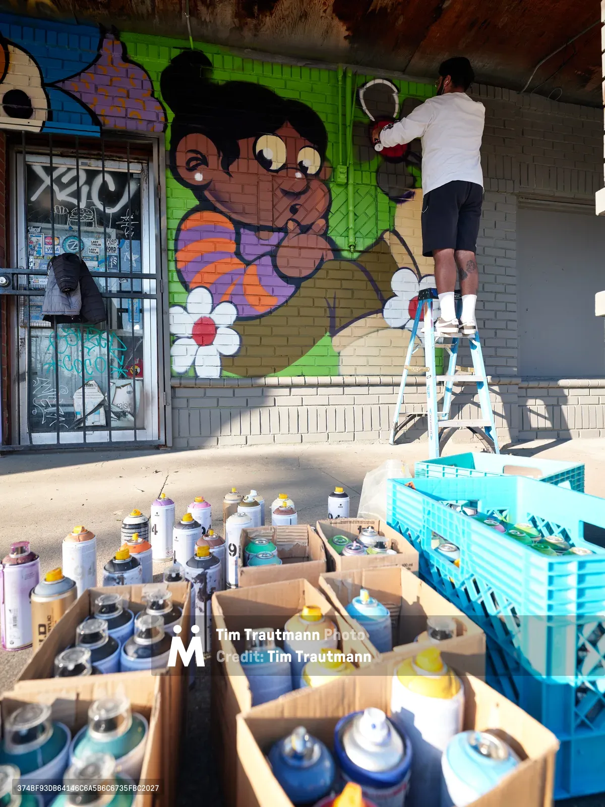 Muralist Heysus works from a ladder to paint a colorful character mural on a brick wall in Portland's Alberta neighborhood. The scene captures the artist mid-creation, surrounded by dozens of organized spray paint cans in cardboard boxes scattered across the concrete sidewalk. Warm afternoon light illuminates the vibrant green and brown tones of the emerging artwork, while a storefront window reflects the creative energy of this Northeast Portland street corner.