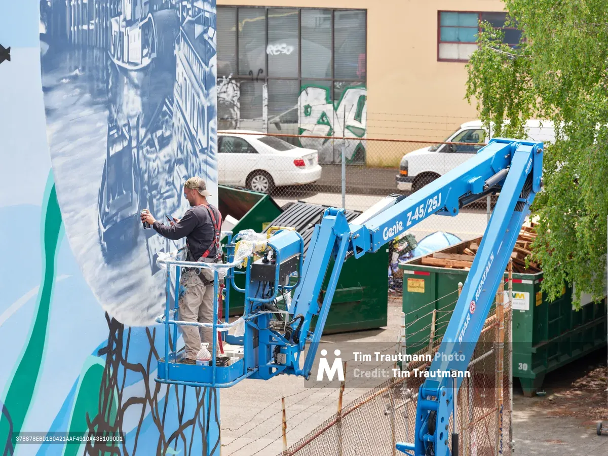 Jeremy Nichols and Devin Finley work methodically from a bright blue Genie lift, applying layers of paint to create a large-scale mural on a weathered building wall in Portland's Central Eastside district. The scene captures the intersection of industrial infrastructure and artistic expression, with green dumpsters and chain-link fencing framing the creative process. Afternoon light illuminates the blue and teal artwork emerging on the concrete surface, while the artists navigate the hydraulic platform with practiced precision.