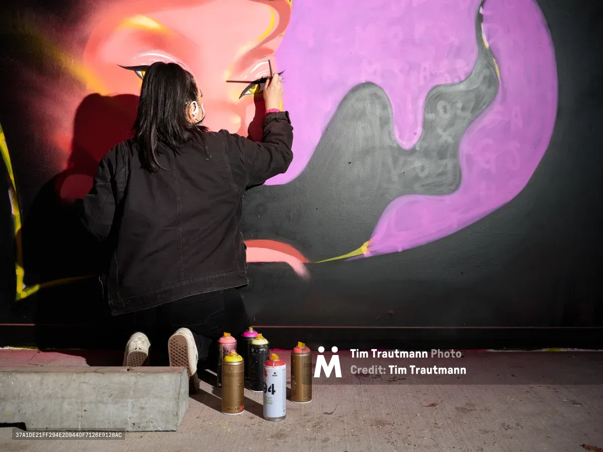 Multidisciplinary artist Vilatose delicately applies finishing touches to an eye detail within a large-scale mural featuring swirling purple and orange forms against a dark background. Working in the intimate confines of Clay Creative's parking garage in Portland's Hosford-Abernethy neighborhood, the artist crouches before their canvas with precise brush strokes while an array of spray paint cans waits nearby on the concrete floor. The dramatic interplay of warm coral highlights and cool violet shadows creates an otherworldly atmosphere in this underground creative sanctuary.