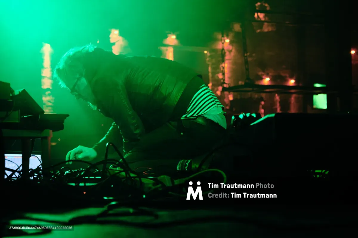 Electronic musician Eli Savage, performing as ellipsism, leans intimately over his synthesizer setup during a hypnotic set at RXFest 2019. Emerald stage lighting bathes the performer and his analog equipment in an otherworldly glow, while the iconic brick interior of Dante's provides a gritty backdrop to the ethereal soundscape. The image captures a moment of pure artistic immersion, as tangled cables and glowing displays create a technological altar of sound in Portland's legendary music venue.