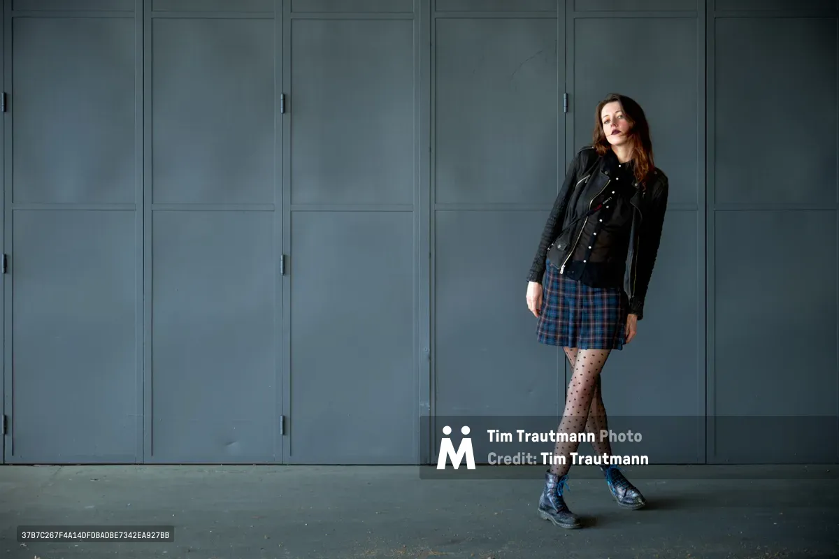 Fashion model Rhianna Atwood leans confidently against weathered steel doors in Portland's Central Eastside industrial district. Dressed in an edgy combination of black leather jacket, plaid mini skirt, and polka dot tights, she embodies contemporary urban style. The muted gray-blue tones of the industrial architecture create a striking minimalist backdrop, with natural light casting subtle shadows across the concrete floor.