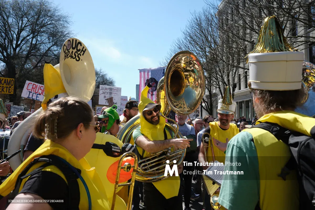 The Unpresidented Brass Band commands attention in Portland's Old Town Historic District during a March 2026 anti-monarchy demonstration. Musicians in vibrant yellow shirts wield gleaming tubas and sousaphones beneath winter-bare trees, their instruments catching the crisp afternoon light. Protest signs declaring "CRUSH ICE" and other political messages create a backdrop of civic unrest, while the band's theatrical energy transforms Southwest Ankeny Street into a stage for musical resistance against perceived authoritarianism.