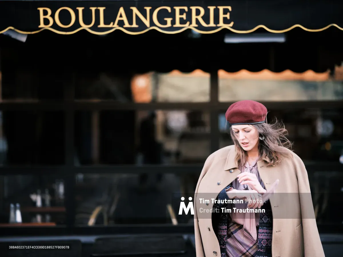 A young woman in a burgundy beret and flowing beige trench coat pauses outside a French bakery on Portland's Alberta Street, her windswept hair catching the soft daylight. The classic black storefront with gold 'BOULANGERIE' lettering creates an atmospheric backdrop, evoking the timeless charm of European café culture transplanted to Oregon's vibrant Vernon neighborhood. Her contemplative downward gaze and elegant layered clothing suggest a quiet moment of urban solitude amid the bustling Northeast Portland streetscape.