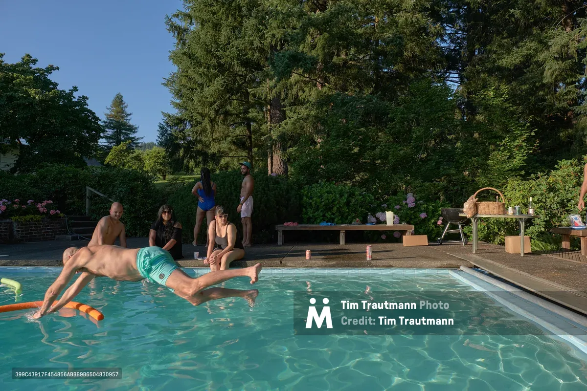 A dynamic moment captures a swimmer mid-dive into crystal-clear pool water while friends gather around the deck in relaxed summer attire. The scene unfolds in a lush orchard setting in Newberg, Oregon, where towering evergreens and blooming hydrangeas create a natural backdrop. Golden afternoon light illuminates the gathering, highlighting the contrast between the active swimmer and the leisurely observers positioned on wooden decking and poolside furniture.