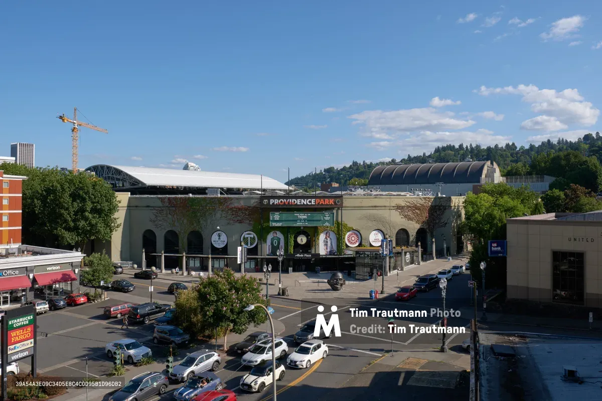 An elevated perspective captures the iconic Providence Park stadium complex in Portland's Goose Hollow neighborhood, where the massive curved venue dominates the urban landscape beneath a brilliant azure sky dotted with wispy clouds. The foreground reveals a bustling commercial intersection with parked cars arranged in neat rows, while construction cranes pierce the skyline beyond the stadium's distinctive architecture. The scene embodies the vibrant confluence of sports, commerce, and urban development in this Pacific Northwest metropolitan hub.
