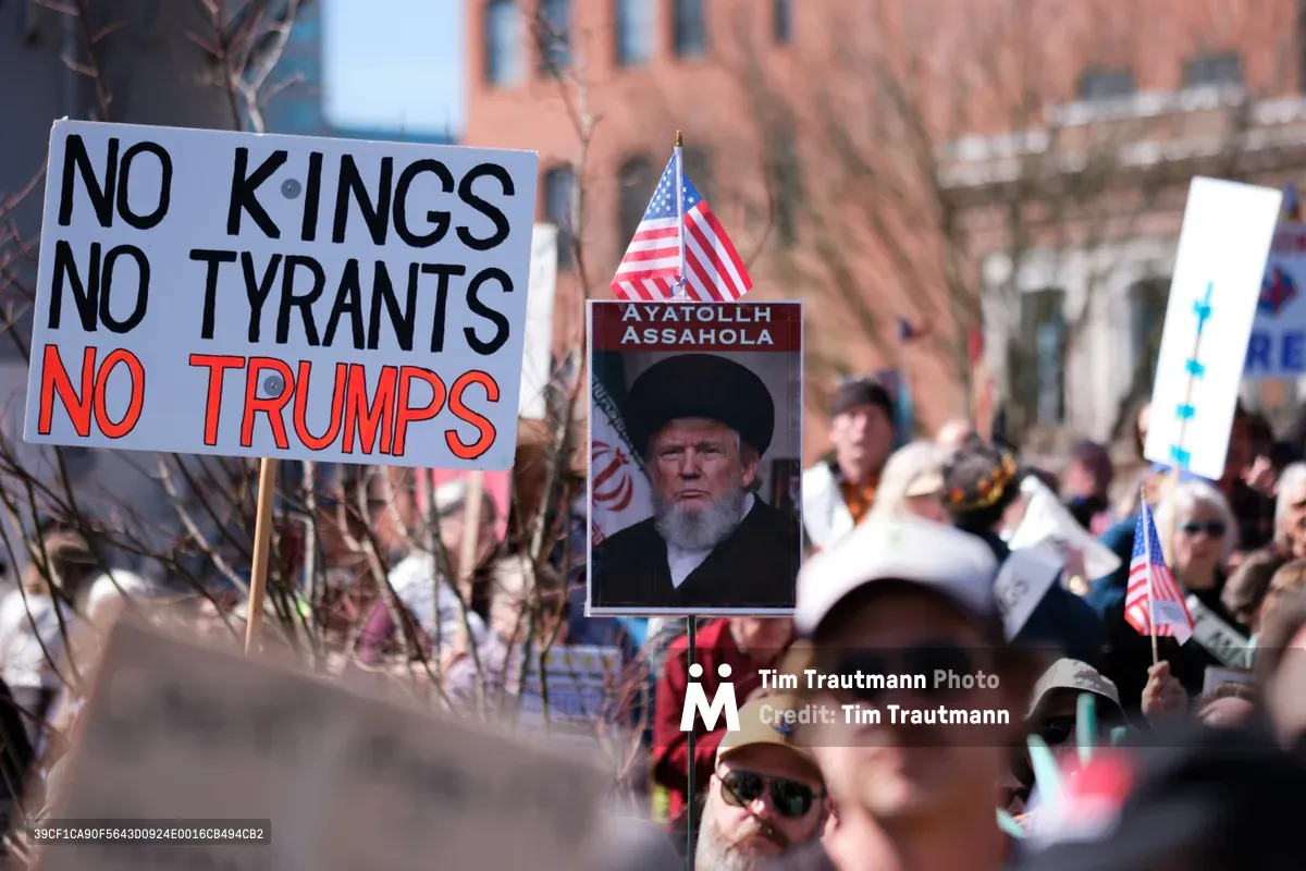 Protesters gather in Portland's historic Skidmore District, wielding handmade signs declaring "NO KINGS NO TYRANTS NO TRUMPS" alongside an altered portrait labeled "Ayatollah Assahola." The afternoon light filters through bare winter branches, casting shadows across the crowd of demonstrators who fill Southwest Ankeny Street. American flags punctuate the scene as citizens exercise their constitutional right to dissent in the heart of Old Town's brick-lined streets.