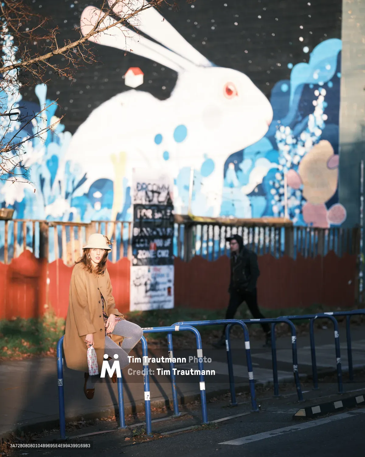 A young woman in a camel coat and knit hat sits pensively on blue metal barriers along Northeast Alberta Street in Portland's vibrant arts district. Behind her, a massive colorful mural featuring a whimsical white rabbit dominates the brick wall, while bare winter branches frame the scene and a figure in dark clothing walks past in the background. The overcast lighting creates a contemplative, urban atmosphere characteristic of Pacific Northwest winters.